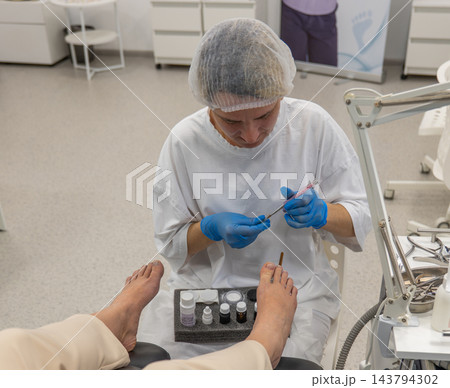 Careful specialist delivers pedicure treatment to person resting feet in chair, inspecting toenail condition while working with fine medical tools during clinical hygiene appointment. 143794302