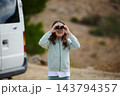 Girl looking through binoculars beside camper van on a family road-trip in a hilly outdoor landscape 143794357