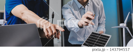 Two Asian engineers wearing hardhats and safety vests discussing solar panel installation and renewable energy project in the office. 143794557