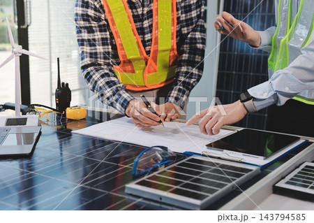 Two Asian engineers wearing hardhats and safety vests discussing solar panel installation and renewable energy project in the office. Two Asian engineers wearing hardhats and safety vests discussing solar panel installation and renewable energy project in the office. 143794585