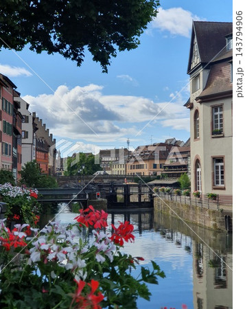Picturesque Petite France canal scene in Strasbourg with colorful flower boxes, historic timbered houses, pedestrian bridge and calm water reflections under a bright blue summer sky. Charming 143794906