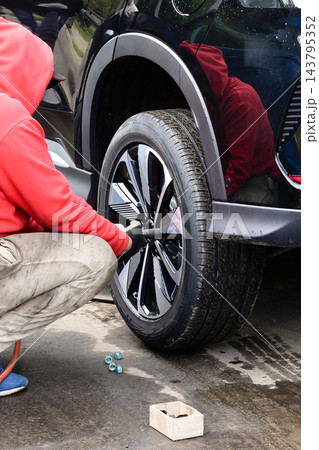 Guy changes wheel on vehicle in a parking area during the day while wearing a red hoodie 143795352