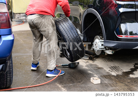 Man changes tire on car in auto repair shop during the day with nearby vehicles parked outside Man changes tire on car in auto repair shop during the day with nearby vehicles parked outside 143795353