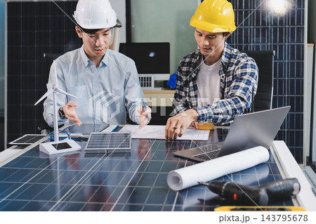 Two Asian engineers wearing hardhats and safety vests discussing solar panel installation and renewable energy project in the office. Two Asian engineers wearing hardhats and safety vests discussing solar panel installation and renewable energy project in the office. 143795678