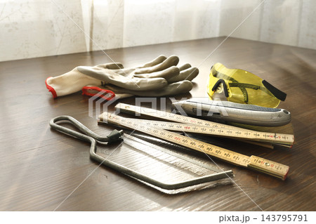 Mini hacksaw folding ruler and protective gear on wooden workshop table. Manual carpentry measuring cutting tools set for home repair. Craftsmanship renovation project building hardware. Mini hacksaw folding ruler and protective gear on wooden workshop table. Manual carpentry measuring cutting tools set for home repair. Craftsmanship renovation project building hardware. 143795791