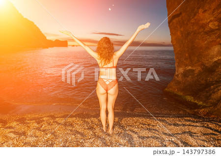 Pebbled beach getaway, woman wearing red lace lingerie stands facing the ocean with her arms raised at sunset Pebbled beach getaway, woman wearing red lace lingerie stands facing the ocean with her arms raised at sunset 143797386