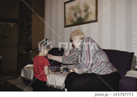 An elderly woman a grandmother brings fruit on a plate to her young grandson in the room and feeds him. Caring for children and the elderly parental love. An elderly woman a grandmother brings fruit on a plate to her young grandson in the room and feeds him. Caring for children and the elderly parental love. 143797771