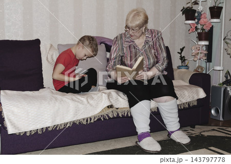 grandmother is reading colorful book grandson, has leaned in closer to listen. grandmother wearing glasses, boy, dressed red T-shirt, is intently examining the book and listening to the story. grandmother is reading colorful book grandson, has leaned in closer to listen. grandmother wearing glasses, boy, dressed red T-shirt, is intently examining the book and listening to the story. 143797778