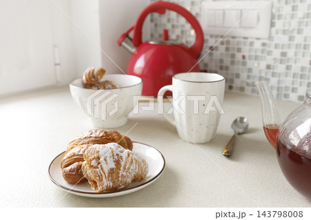 Fresh croissants on plate with hot fruit tea mug and red kettle. Morning breakfast cozy kitchen tea break sweet bakery snack. French pastry domestic brunch cafe lifestyle dining banner. Fresh croissants on plate with hot fruit tea mug and red kettle. Morning breakfast cozy kitchen tea break sweet bakery snack. French pastry domestic brunch cafe lifestyle dining banner. 143798008