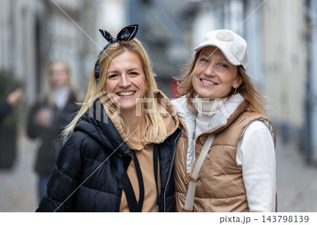 Two happy women laugh together while posing on cobblestone street, wearing jackets during autumn city walk. Joyful friendship portrait captures spontaneous happiness, travel memories and connection. 143798139