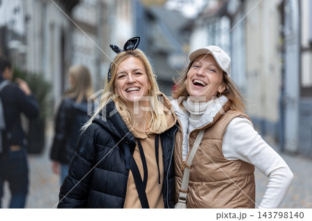 Two happy women laugh together while posing on cobblestone street, wearing jackets during autumn city walk. Joyful friendship portrait captures spontaneous happiness, travel memories and connection. 143798140