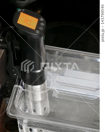 Sous vide immersion circulator heating water in a transparent plastic container. Cooking equipment used for precision temperature control in modern culinary arts and home food preparation. Sous vide immersion circulator heating water in a transparent plastic container. Cooking equipment used for precision temperature control in modern culinary arts and home food preparation. 143799540