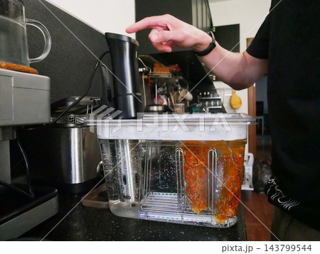 Man starting sous vide immersion circulator to cook food in water bath. Chef preparing meal using precision temperature cooking technique in home kitchen. Culinary technology equipment. Man starting sous vide immersion circulator to cook food in water bath. Chef preparing meal using precision temperature cooking technique in home kitchen. Culinary technology equipment. 143799544