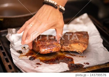 Hand lifting a piece of glazed grilled meat rib with visible exposed bones resting on parchment paper over a baking tray. Hand lifting a piece of glazed grilled meat rib with visible exposed bones resting on parchment paper over a baking tray. 143799576