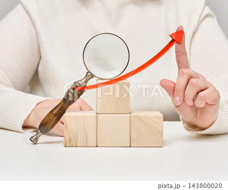 Woman's hand pointing at a red growth arrow above a magnifying glass and wooden cubes 143800020
