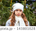 Sad girl in white hat and scarf standing by a decorated Christmas tree outdoors in winter 143801028