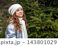 Smiling girl in white hat and scarf standing by an evergreen Christmas tree outdoors in winter 143801029