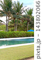 Tropical garden surrounds swimming pool with tall palms and clipped hedges. Landscaped grounds include blue sun umbrella, bright lawn and clear turquoise water in calm resort setting. 143802066