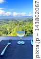Glass of water rests on outdoor dining table overlooking green hills. Clear drink, fork and knife set tranquil place setting beneath bright blue sky and scattered clouds. Glass stands beside cutlery. 143802067