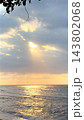 Coastal sunset illuminates rippling ocean water beneath wide cloud filled sky. Golden reflections lead toward horizon, while silhouetted leaves frame tranquil destination for tourism. 143802068