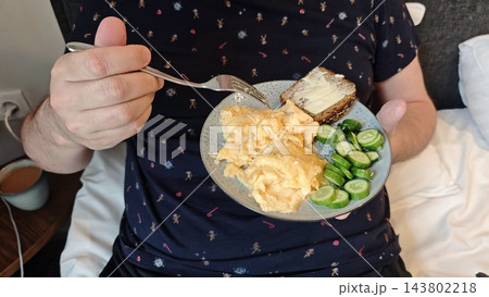 Person holding a plate of scrambled eggs, sliced cucumber, and toast while seated indoors, viewed from above in a casual breakfast scene Person holding a plate of scrambled eggs, sliced cucumber, and toast while seated indoors, viewed from above in a casual breakfast scene 143802218