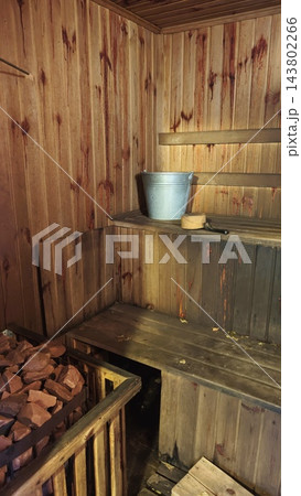 Rustic wooden sauna interior featuring a bench, metal bucket, ladle, and heated stones beside a dark plank wall and floor Rustic wooden sauna interior featuring a bench, metal bucket, ladle, and heated stones beside a dark plank wall and floor 143802266