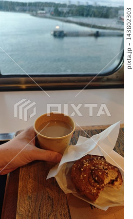 A hand holds coffee beside a glazed pastry on a wooden table, with calm water, a dock, and distant shoreline visible through the ferry window A hand holds coffee beside a glazed pastry on a wooden table, with calm water, a dock, and distant shoreline visible through the ferry window 143802303