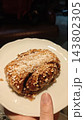 Cinnamon bun covered with pearl sugar and cinnamon on a white scalloped plate, held by a hand in a dim indoor setting 143802305