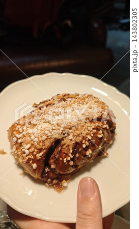 Cinnamon bun covered with pearl sugar and cinnamon on a white scalloped plate, held by a hand in a dim indoor setting 143802305