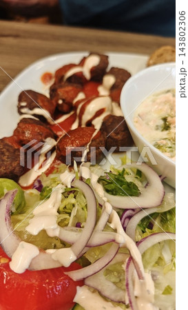 Meatballs with creamy sauce, salad, tomato, and dip arranged on a white plate over a wooden table, close up of a plated meal 143802306