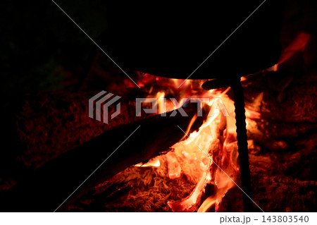 Traditional cooking pot silhouetted above burning firewood at night, glowing flames and embers beneath outdoor cookware for rustic campfire cooking, survival, warmth and rural lifestyle Traditional cooking pot silhouetted above burning firewood at night, glowing flames and embers beneath outdoor cookware for rustic campfire cooking, survival, warmth and rural lifestyle 143803540