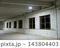 A concrete parking garage wall has repeating windows, overhead lights, and yellow pavement markings. The long interior scene shows pipes, signs, reflections, and an empty urban parking area. 143804403
