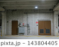 A concrete parking garage wall shows brown utility doors, electrical meters, pipes, and overhead lights. The industrial scene includes block walls, yellow pavement markings, signs, and structural 143804405