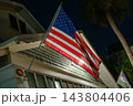 An American flag hangs from a building facade at night under bright exterior lighting. The patriotic scene shows red stripes, blue stars, a roofline, windows, and palm trees in the background. 143804406