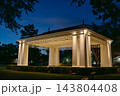 A white park pavilion glows with warm lights under a deep blue evening sky. Trees, hedges, benches, and architectural columns surround the peaceful outdoor structure. 143804408