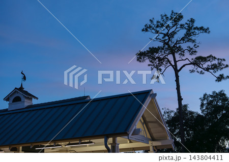 A Winter Park station roof sits below a tall tree silhouette in the blue evening sky. The quiet architectural detail includes warm station lights, treetops, and soft twilight color. A Winter Park station roof sits below a tall tree silhouette in the blue evening sky. The quiet architectural detail includes warm station lights, treetops, and soft twilight color. 143804411