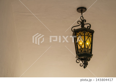 A vintage hanging lantern glows with warm light against a plain ceiling. The ornate metal frame and soft shadows create a classic architectural detail. 143804417