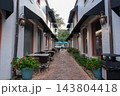 An empty cafe courtyard has brick pavement, outdoor tables, potted flowers, white buildings, and black awnings. The narrow walkway, lanterns, blue umbrellas, and shop windows create a quiet shopping 143804418