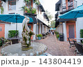 A courtyard fountain stands beside blue umbrellas, cafe tables, white buildings, and brick pavement. The flowers, balconies, black awnings, outdoor chairs, and pedestrian walkway create a charming 143804419