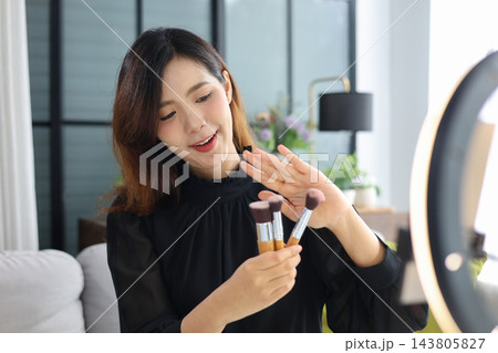 Female content creator filming beauty vlog holding cosmetics brushes for social media video production. 143805827