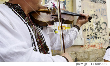 Adult man wearing a traditional embroidered vyshyvanka plays a vintage violin on city street. The scene emphasizes cultural identity, heritage and the expressive character of live music. Slow mo Adult man wearing a traditional embroidered vyshyvanka plays a vintage violin on city street. The scene emphasizes cultural identity, heritage and the expressive character of live music. Slow mo 143805859