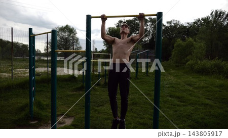 Muscular athlete doing pull ups on horizontal bar at sports ground. Young shirtless sportsman exercising at nature. Strong sporty man training outdoor. Concept of healthy and active lifestyle. Slow mo Muscular athlete doing pull ups on horizontal bar at sports ground. Young shirtless sportsman exercising at nature. Strong sporty man training outdoor. Concept of healthy and active lifestyle. Slow mo 143805917