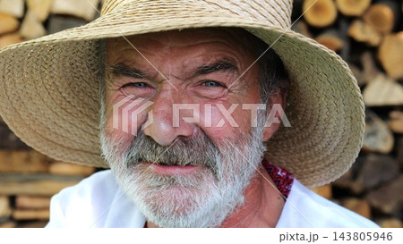 Older smiling male wearing an embroidered shirt looking into camera outdoor. Portrait of senior ukrainian man in straw hat against background of woodshed. Cultural heritage and ethnic roots concept 143805946