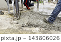 Male hands in protective gloves uses trowel to spread and level fresh cement in yard. Another worker distributes and moves the mixture with a shovel preparing the material during construction process 143806026