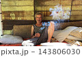 Portrait of homeless adult man sitting on worn couch placed and smoking outdoor. The setting underscores neglect and impermanence, presenting a raw and unfiltered glimpse of difficult living situation 143806030