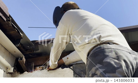 Manual laborer in protective mask welds iron detail at construction. Professional young repairman with welding machine working at the site. Male welder operating with metal on house. Close up 143806033
