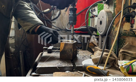 Male hands in protective gloves of mechanic operate a drill press while machining a metal detail at garage. Repairman works carefully in a small workshop, concentrating on technical repair. Close up 143806035