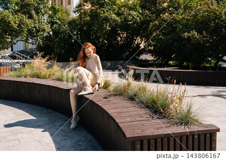 Wide shot of young woman with long red hair sitting on curved wooden bench in city park, tying sneakers. Casual lifestyle moment on sunny summer day. Bright natural light creating strong shadows. Wide shot of young woman with long red hair sitting on curved wooden bench in city park, tying sneakers. Casual lifestyle moment on sunny summer day. Bright natural light creating strong shadows. 143806167