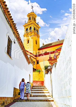 Two women walk up the street leading to the Church of Santiago in Alcala de Guadaira, with the church's bell tower in the background; Seville. 143806310