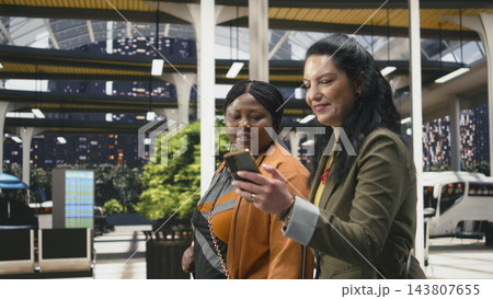 Smartphone, travelers and bus terminal as woman in green blazer checks her mobile phone while companion looks on. Navigation, coordination and digital travel aid shared journey planning at station. Smartphone, travelers and bus terminal as woman in green blazer checks her mobile phone while companion looks on. Navigation, coordination and digital travel aid shared journey planning at station. 143807655